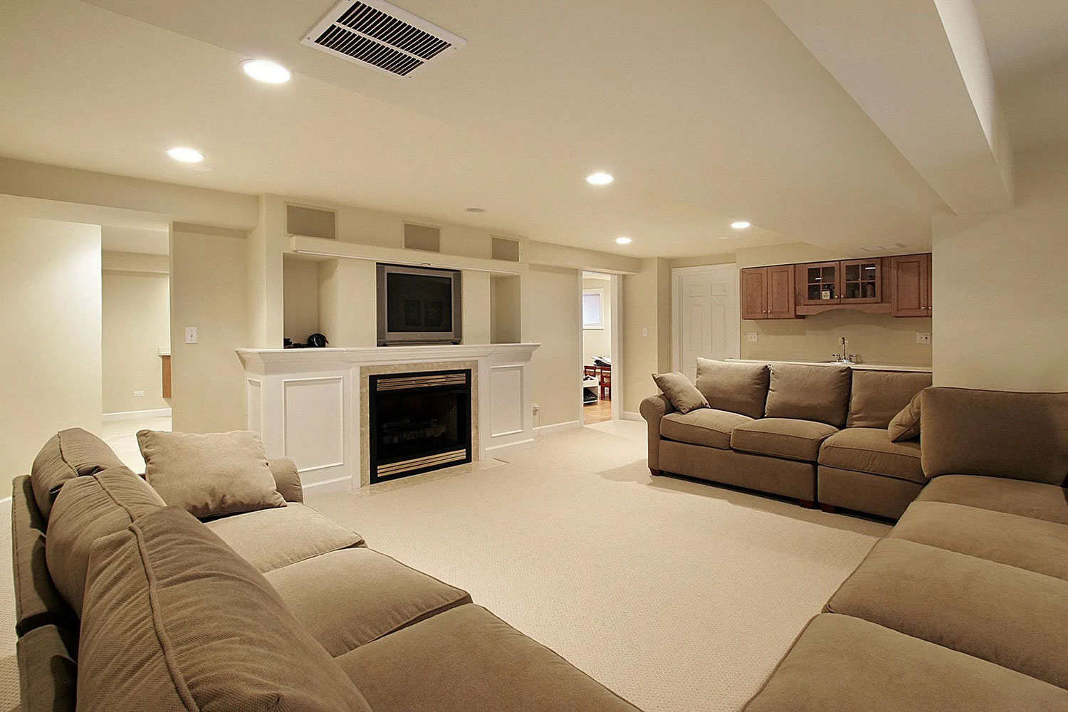 Spacious finished basement living room with beige sofas, a carpeted floor, fireplace, built-in TV shelving, and a compact kitchen area.